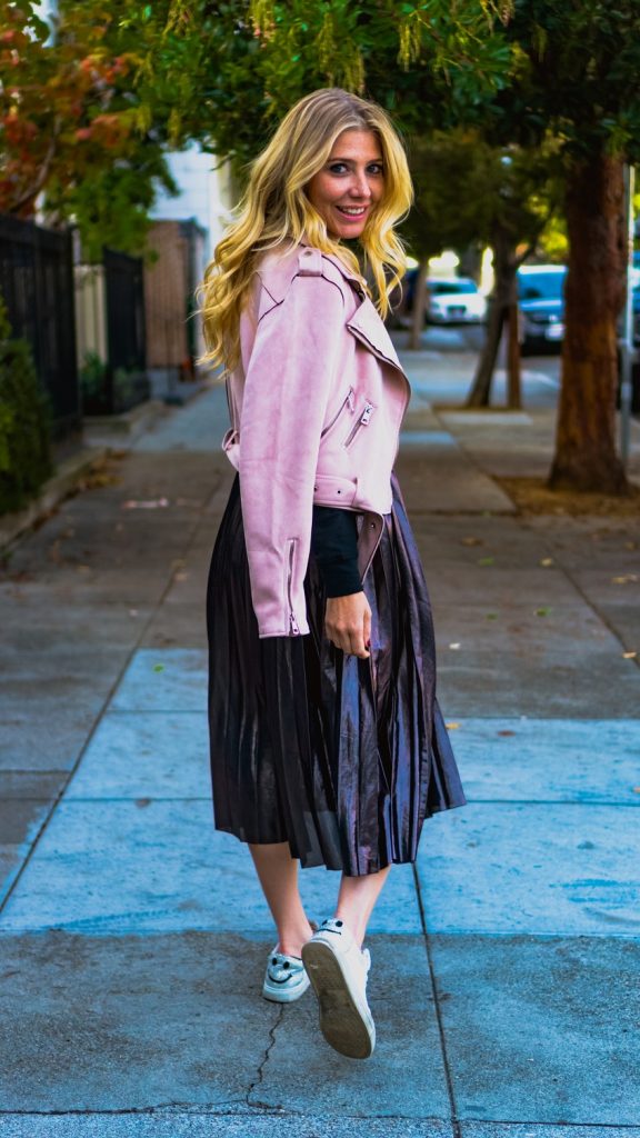 picture of a woman walking on a sidewalk wearing leather coat, skirt and sneakers 